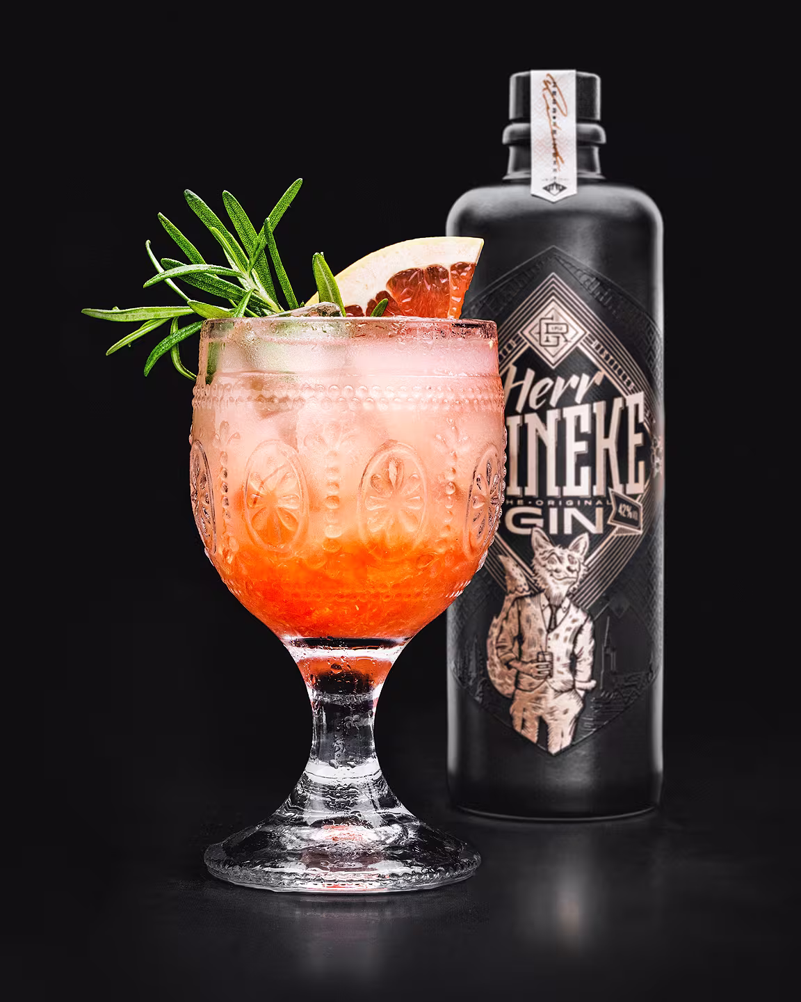 Cocktailglas mit pinkem Drink, dekoriert mit Rosmarin und Grapefruit, vor der mattschwarzen Herr Reineke Gin-Flasche