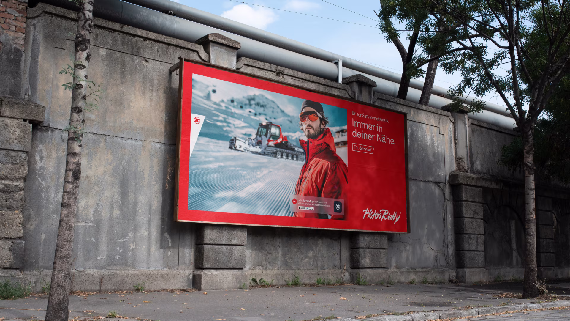 PistenBully-Werbeplakat mit Fahrer vor verschneiter Bergkulisse an einer Betonwand