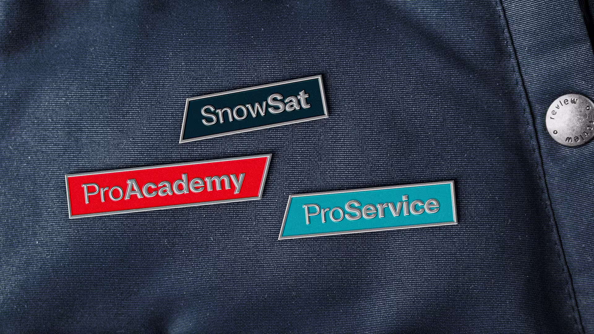 Drei metallische Pins mit den Aufschriften SnowSat, ProAcademy und K-Service auf dunklem Stoff.